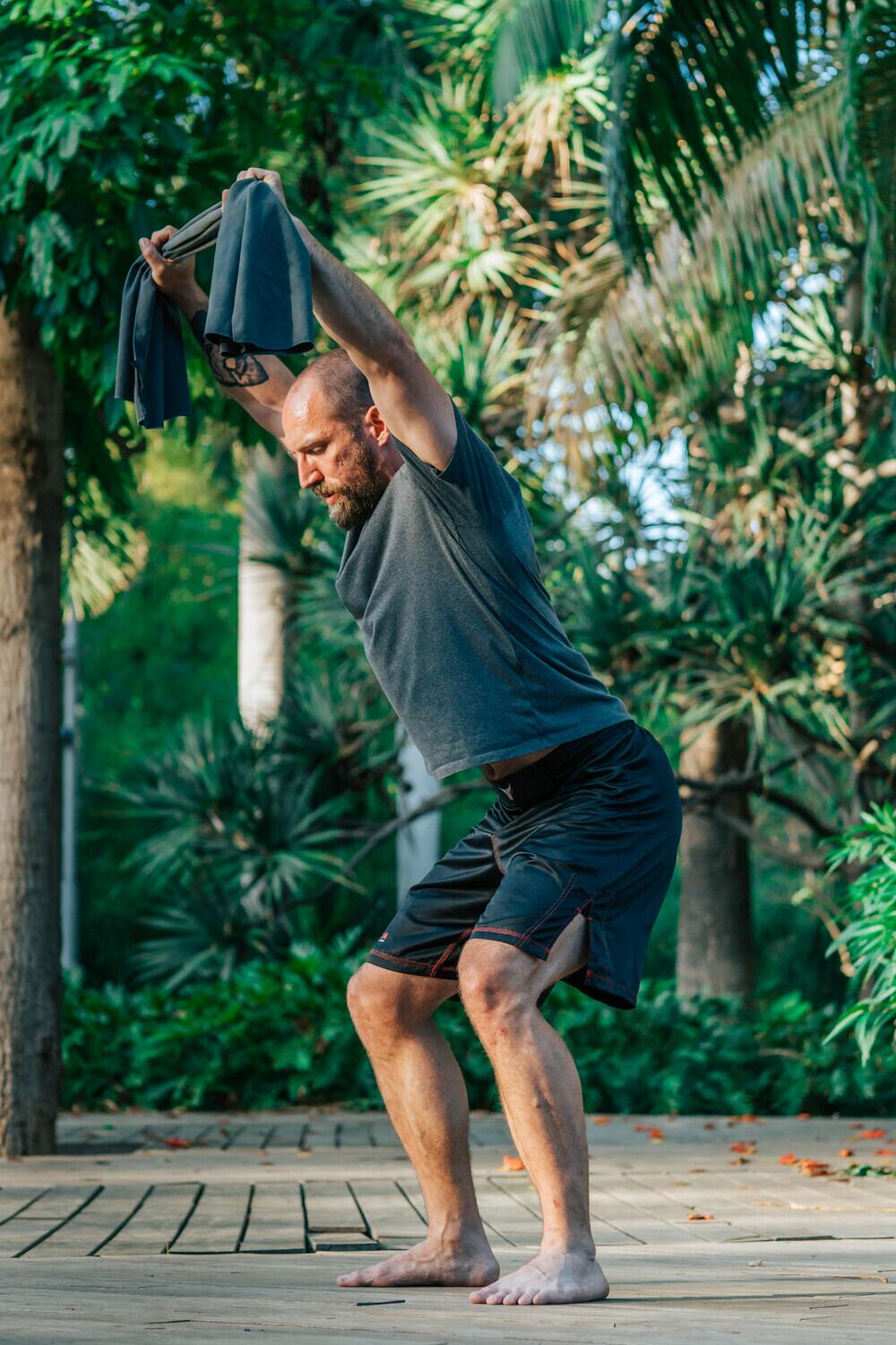 Joe performing an overhead row with a rolled towel, arms extended above his head, training outdoors