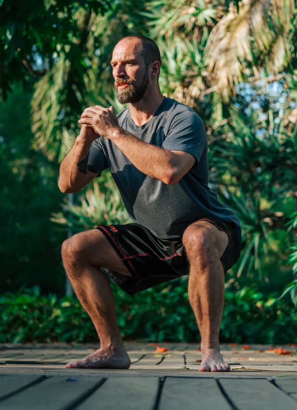 Joe holding the bottom of a deep bodyweight squat, hands clasped at his chest, training outdoors
